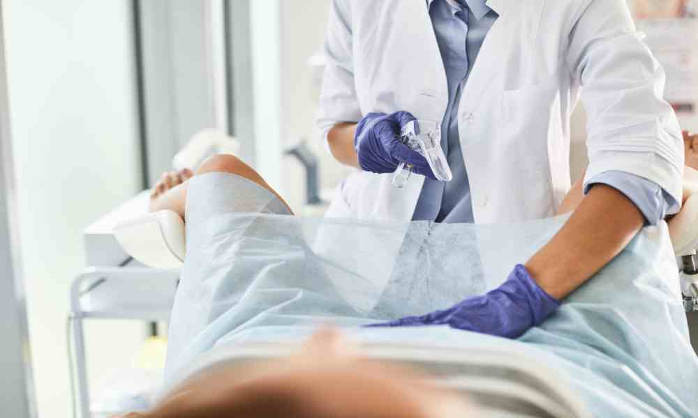 A gynecologist wearing purple gloves and a white lab coat. The doctor is performing a pelvic exam on a patient.