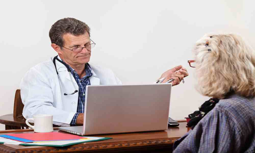 A doctor wearing wireframed glasses, a white lab coat, and a stethoscope looking disapprovingly at a female patient.