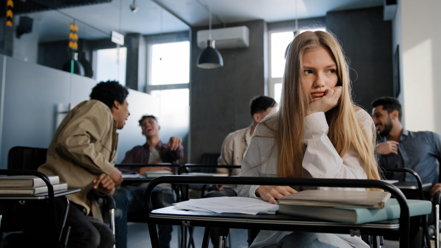 A young woman in a classroom who looks depressed staring off at nothing with other students laughing in the background