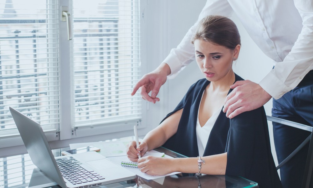 A woman sitting at her desk looking uncomfortable while a man approaches her from behind with his hand on her shoulder.