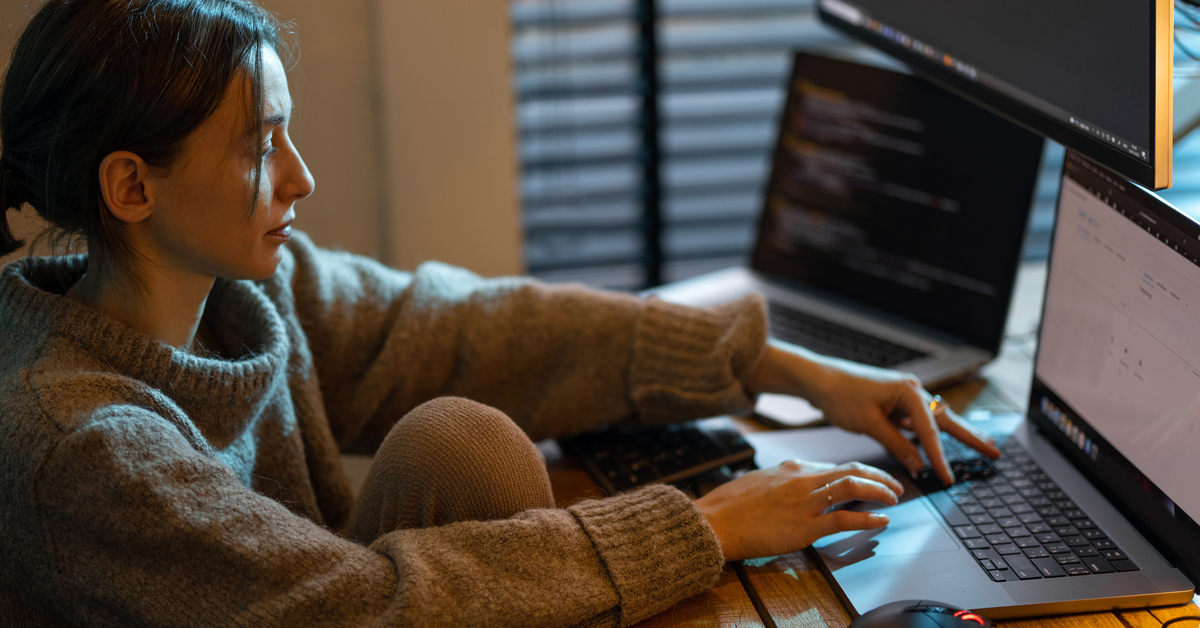 A tired-looking woman wearing a turtle neck sitting at her home work desk in front of two computer screens.