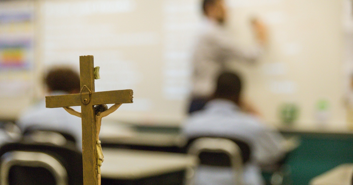 A blurred background of a teacher at the front of a classroom writing on a white board with a crucifix in the foreground.