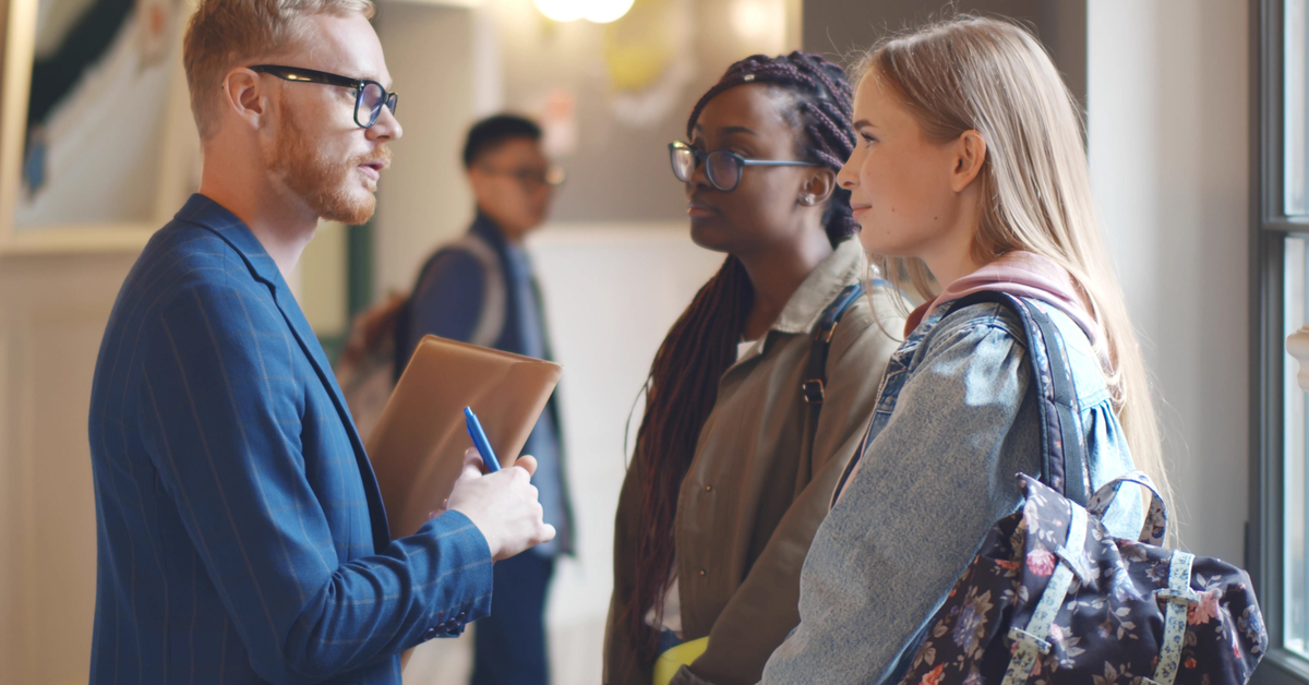 A young male professor wearing glasses and holding a folder talking to two young female students in the hallway.