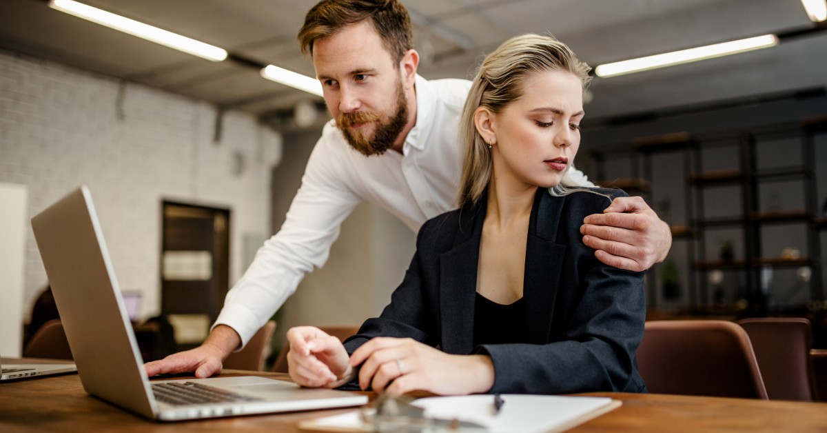 A blonde businesswoman dressed in black sitting at her desk as she looks at the hand of her male coworker on her shoulder.
