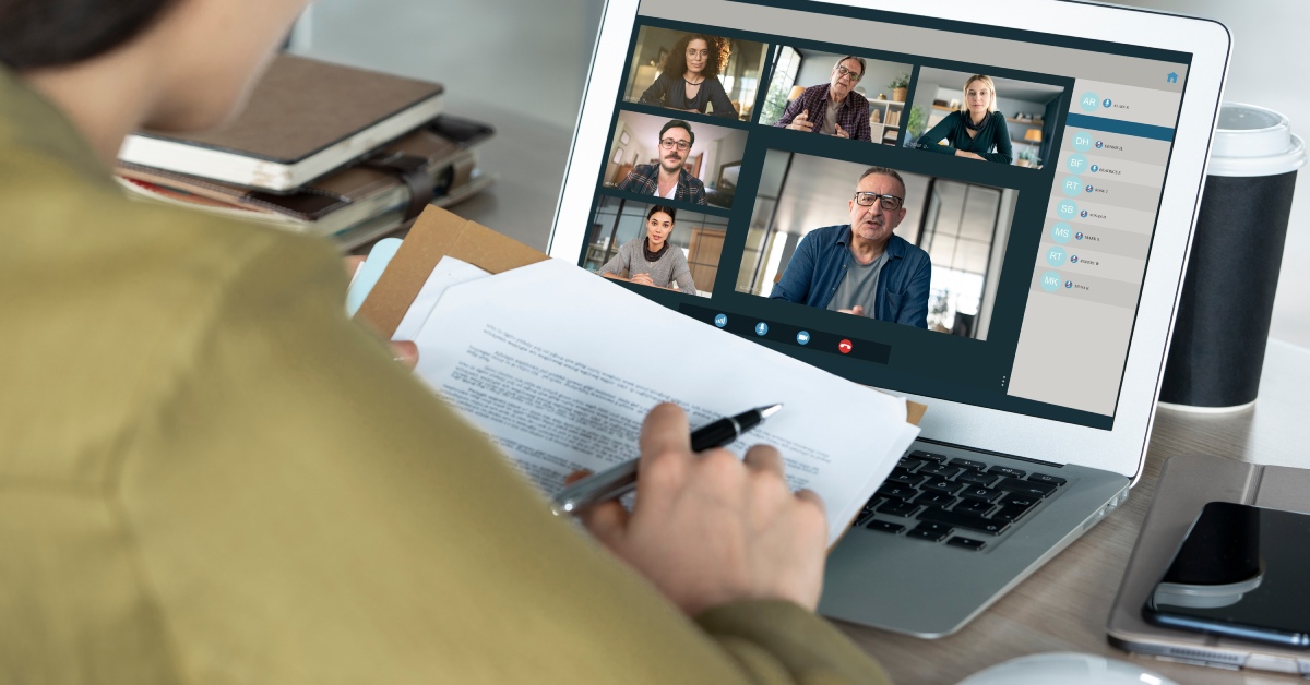 Over the shoulder of a woman looking at a laptop with the computer screen displaying six people on a virtual call.