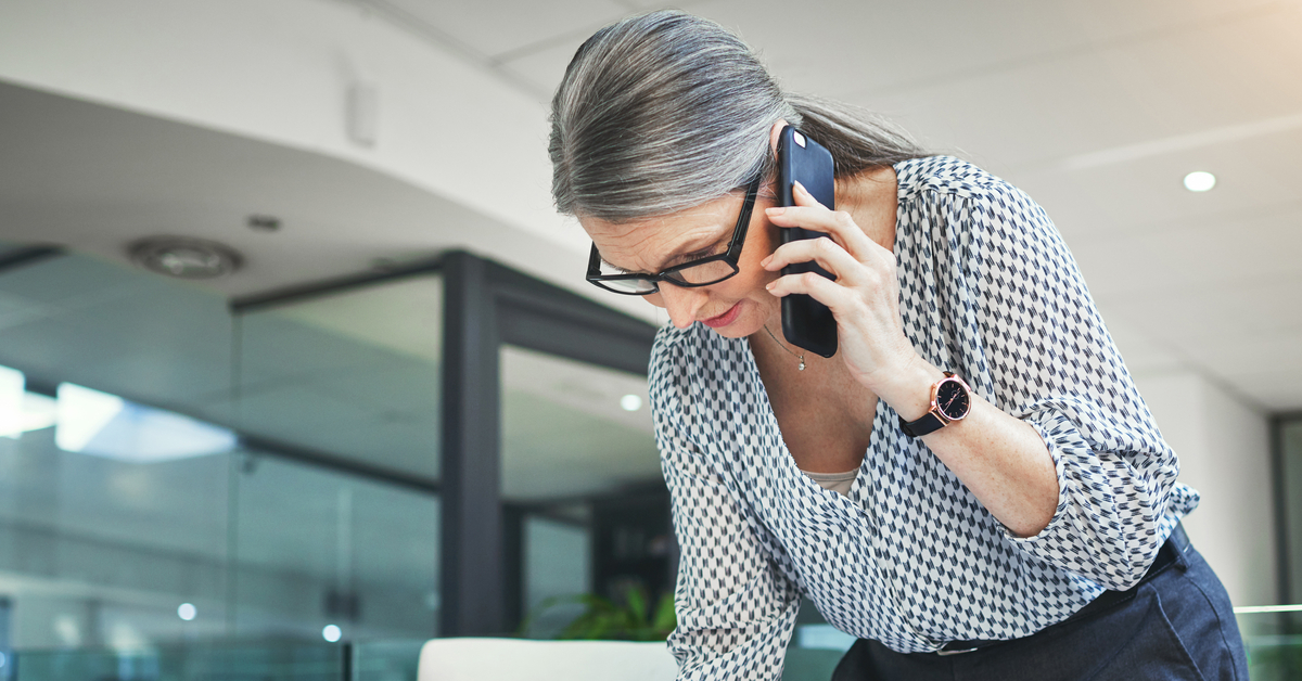 An older woman in glasses, holding up her smartphone to her ear as she writes on her clipboard.