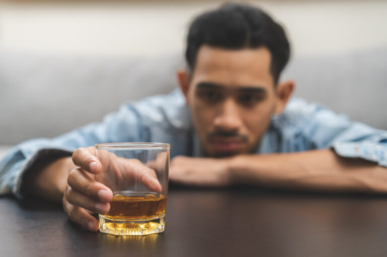 A glass of brown liquor is in the foreground, with a young man reaching out to hold it. The man is blurred in the background.