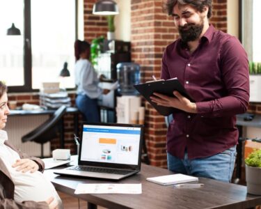 A pregnant woman at her computer desk looking up at the bearded man across from her as he writes on a clipboard.
