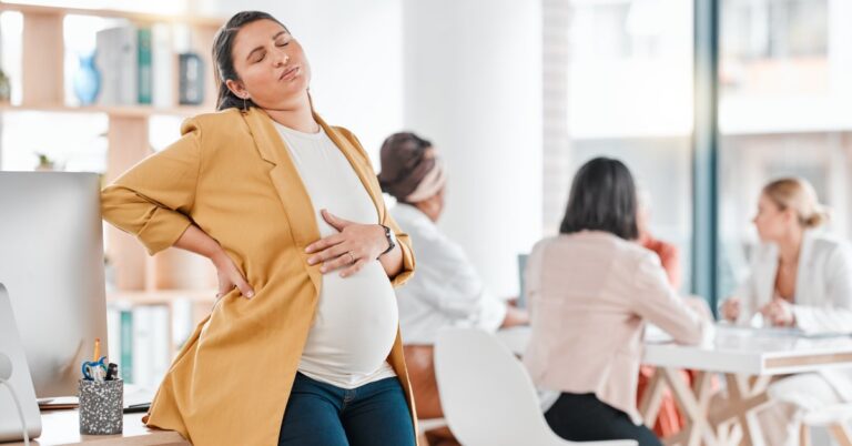 A pregnant woman holding her stomach as she leans back against a desk. There are other women at a table in the background.