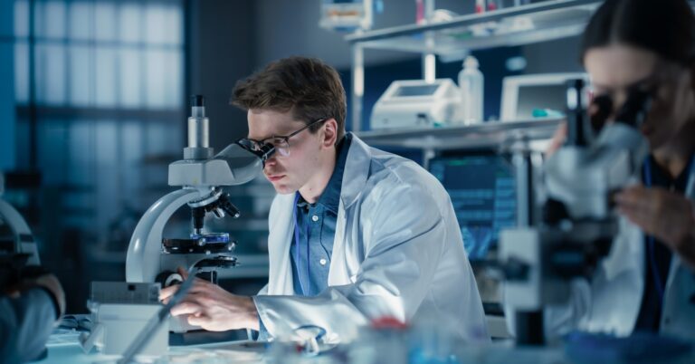A young man in a lab is wearing glasses and a lab coat while looking into the lens of a microscope.