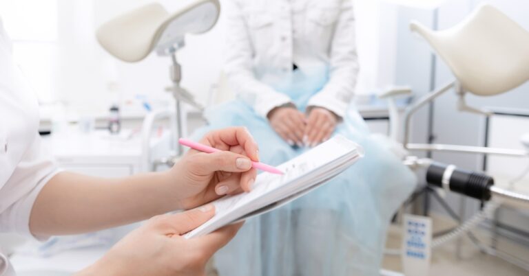A person wearing a medical gown is sits on an exam table. In the foreground, a pair of hands holds a notebook and a pink pen.