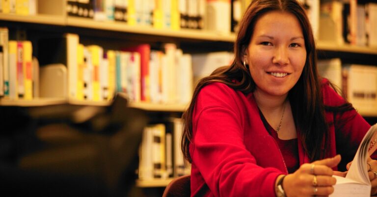 A young woman with long black hair wearing a red cardigan sits at a library table with an open book.