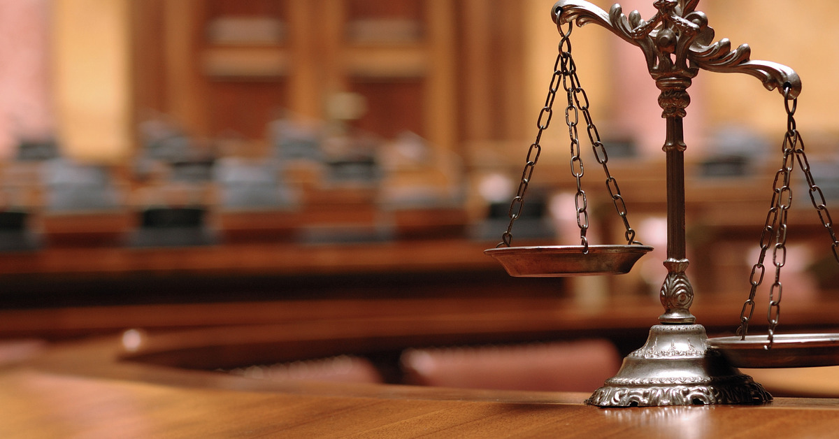 A focus shot of the scales of justice on a wooden table with a courtroom blurred in the background.