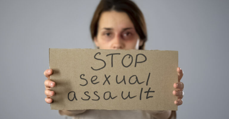 A brunette woman with bruised under-eyes is holding up a cardboard sign that says