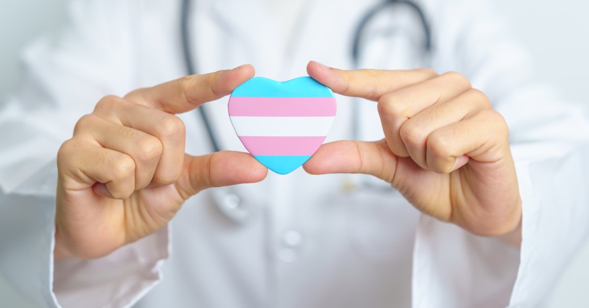 A person wearing a lab coat and a telescope is holding up a heart with the transgender pride flag.