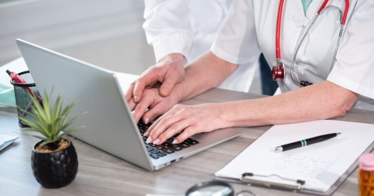 A focus shot of a woman's hands on her keyboard with another hand caressing hers. She is wearing a red stethoscope.