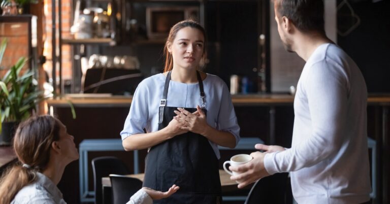 A female server wearing a dark apron is standing between a man and a woman, gesturing angrily at her.
