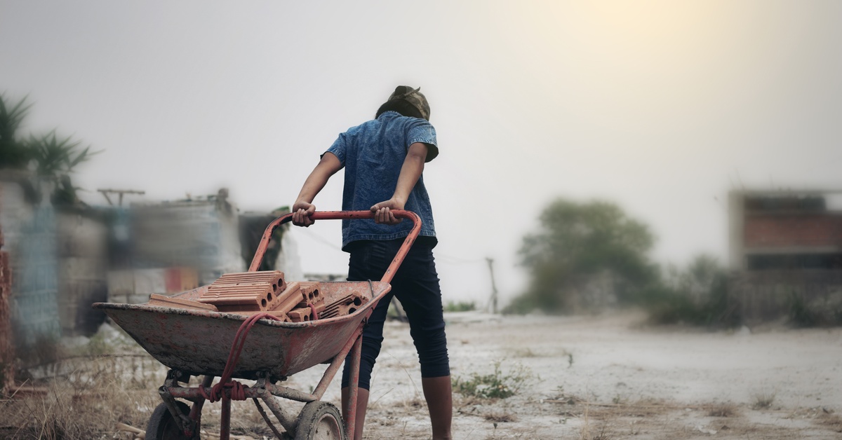 A young person wearing no shoes, dragging a rusty red wheelbarrow full of red bricks through the dirt.