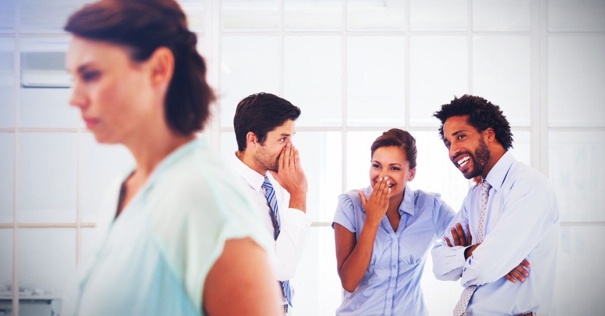 A group of three professionally dressed people laughing and whispering. A woman walks by in front of them.