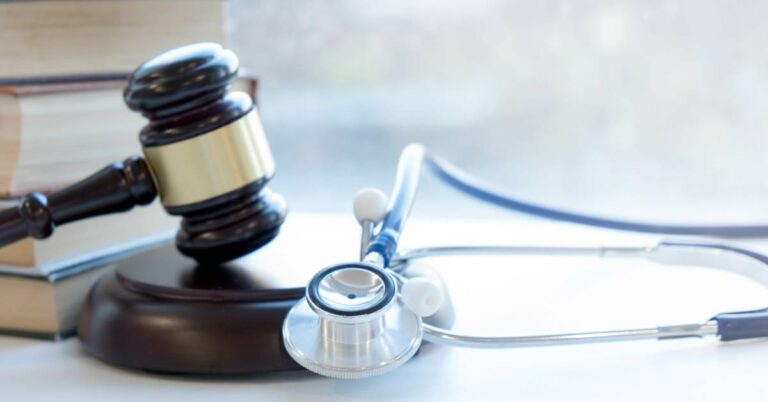 A wooden gavel and gavel stand sat between a stack of four books and a stethoscope, all on a white desk.