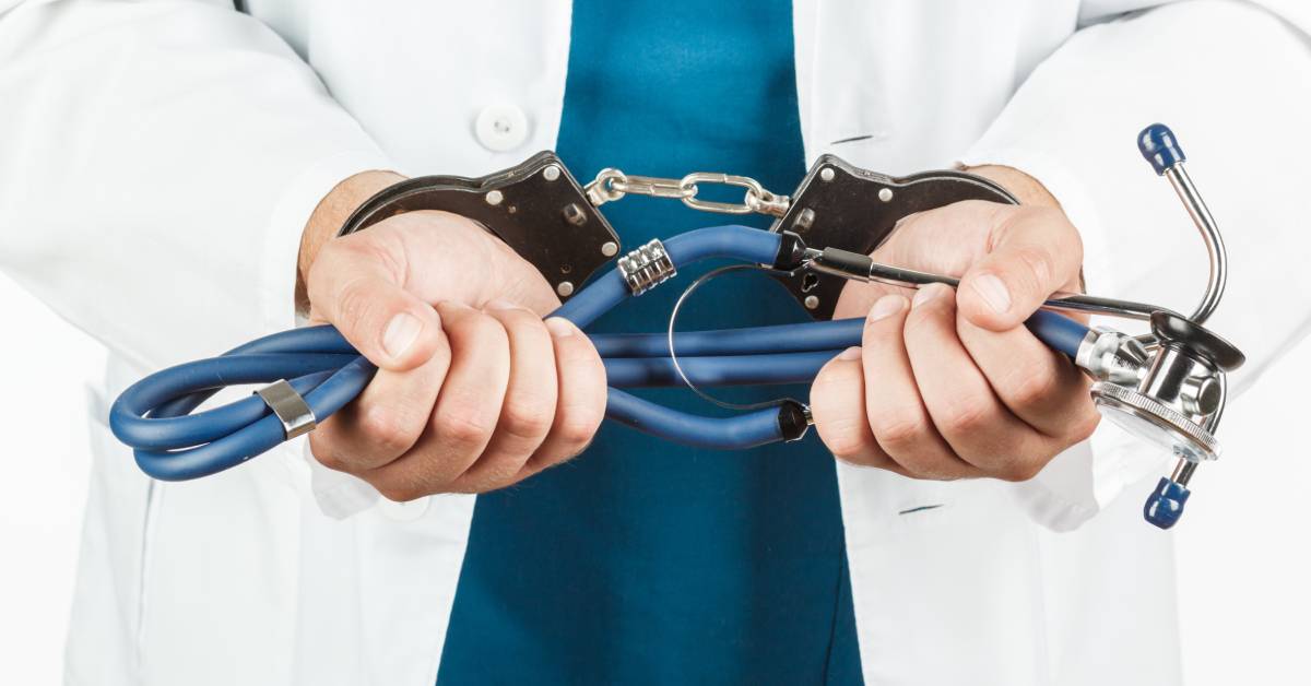 A focus shot of a pair of hands shackled with black handcuffs while holding a folded-up stethoscope.