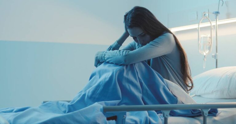 A young woman sitting on a hospital bed in a dimly lit room, holding her head with her knees to her chest.