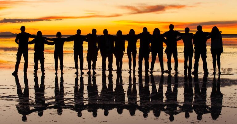 A large group of friends in a line, standing on the beach in front of the sunset; the angle of the light is silhouetting them.