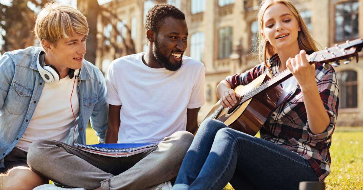 Two young men and a young woman sitting on the lawn of a university, while the girl is playing a guitar.