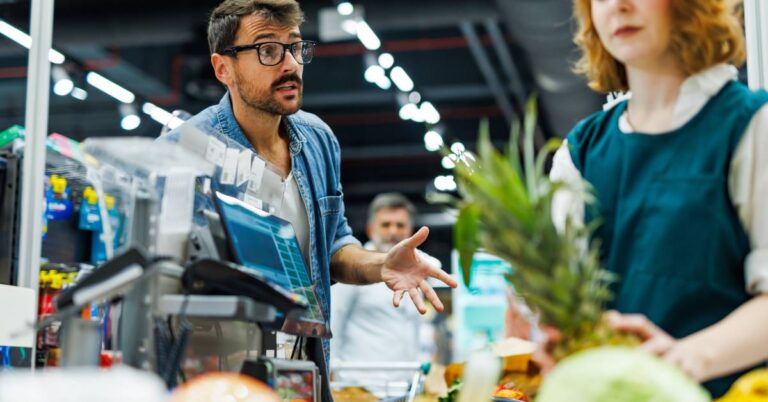 A customer with a beard and glasses is angrily gesturing over the conveyor belt at a grocery store clerk.