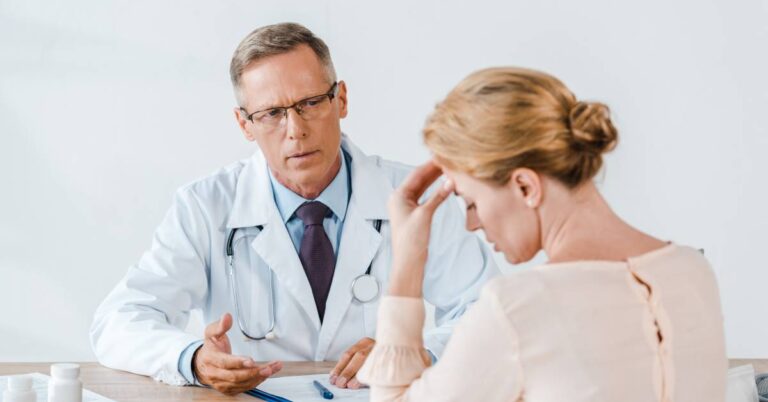 A doctor gesturing to the blonde woman sitting across from him at a desk. She is upset and holding her head in her hand.