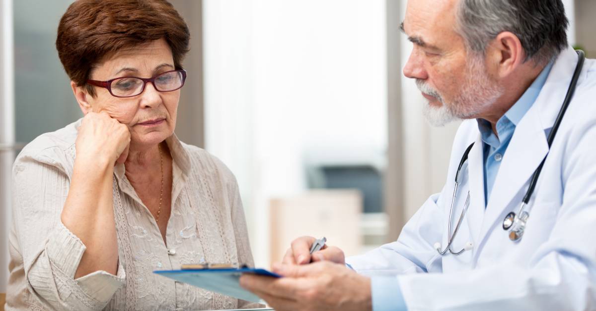 An older woman wearing glasses leans her head against her fist as she looks down at the clipboard her doctor is holding.