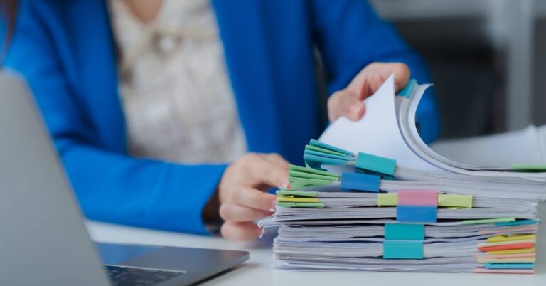 A focus shot of a large set of papers, each with a different sticky note. A woman is shuffling through the papers in the background.