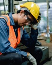 A tired worker is resting his hand on his head. He is wearing an orange high-visibility vest and a yellow hard hat.