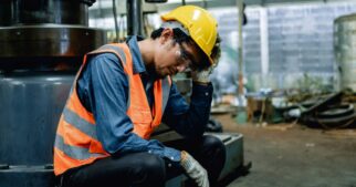 A tired worker is resting his hand on his head. He is wearing an orange high-visibility vest and a yellow hard hat.