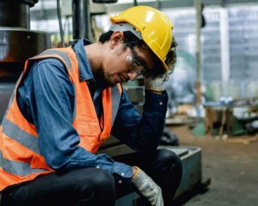 A tired worker is resting his hand on his head. He is wearing an orange high-visibility vest and a yellow hard hat.