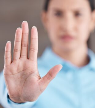 A close-up, focus shot of a woman's hand as she sticks it outward, palm facing the camera; she is blurred out in the background.
