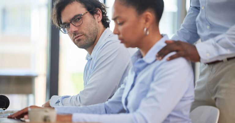 A man sitting next to his female coworker, looking at her face as a pair of hands comfortably massage her shoulders.