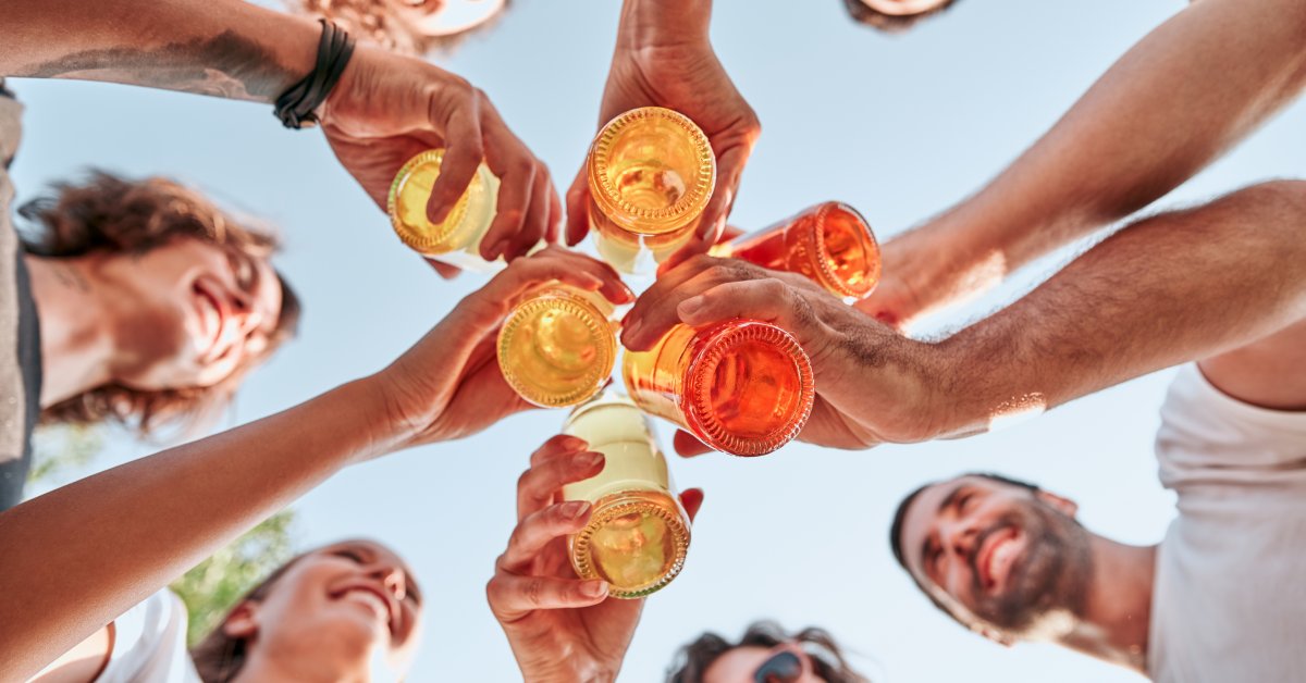 A worm's-eye view of a group of smiling people, standing in a circle, cheering their drinks.