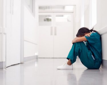 A woman in scrubs sitting on the floor of a clean, white hallway, her knees pulled to her chest and her head resting on them.