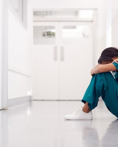 A woman in scrubs sitting on the floor of a clean, white hallway, her knees pulled to her chest and her head resting on them.