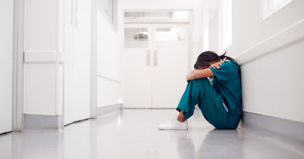 A woman in scrubs sitting on the floor of a clean, white hallway, her knees pulled to her chest and her head resting on them.