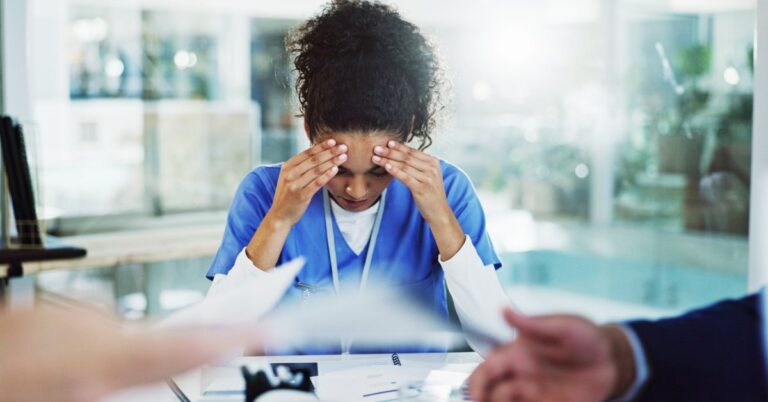 A woman sitting at a table covered in paperwork, holding her forehead, while two other people gesture over the documents.