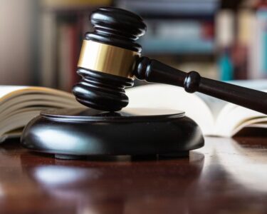 A dark, wooden gavel on top of a wooden block of the same color and material, next to several open books on a table.