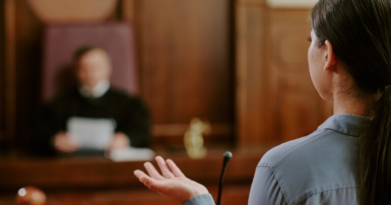 A woman in a courtroom standing before a judge, who's holding a piece of paper. She talks into a tall, skinny microphone.
