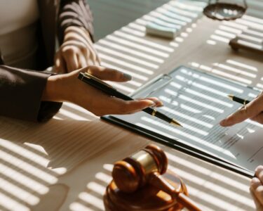 Two people sitting at a desk that has a gavel and a scale of justice on it, holding pencils and pointing at a paper.