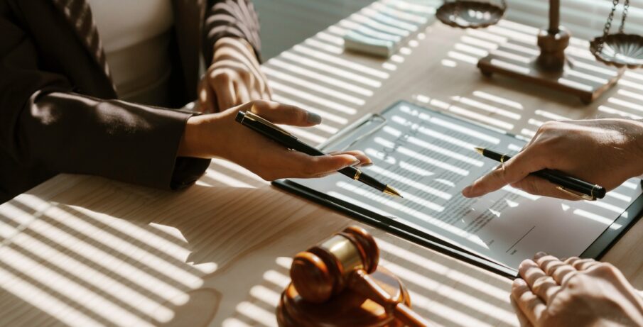 Two people sitting at a desk that has a gavel and a scale of justice on it, holding pencils and pointing at a paper.