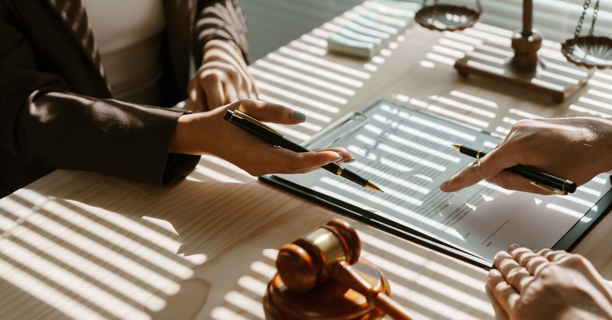 Two people sitting at a desk that has a gavel and a scale of justice on it, holding pencils and pointing at a paper.