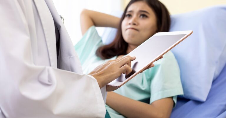 A person in a white coat holding a tablet standing next to a woman in a hospital gown who's lying on an exam bed.