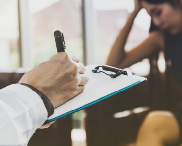 A woman sitting in a chair, resting her head on her hand, while another person in front of her takes notes on a clipboard.