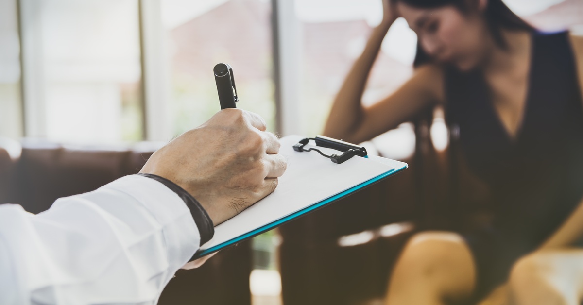A woman sitting in a chair, resting her head on her hand, while another person in front of her takes notes on a clipboard.
